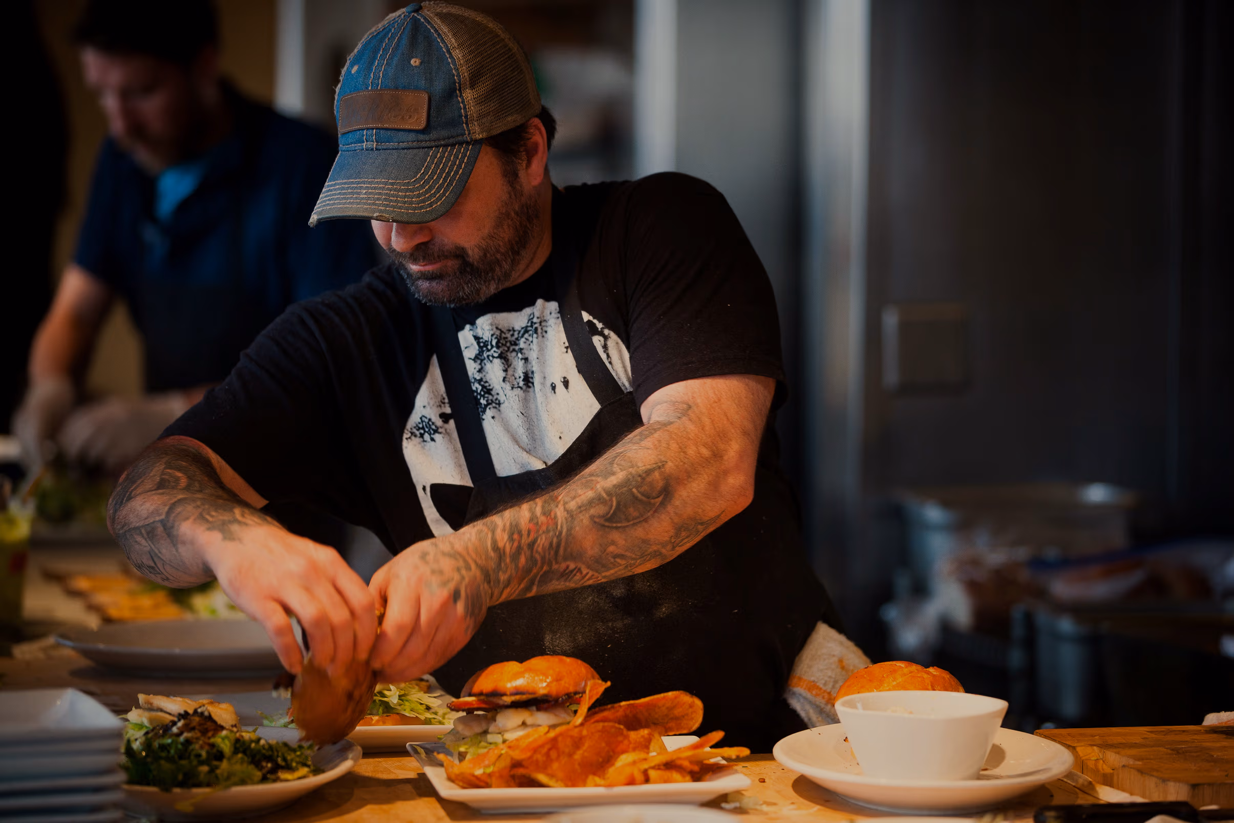 A cook in a cap building a burger at the pass.