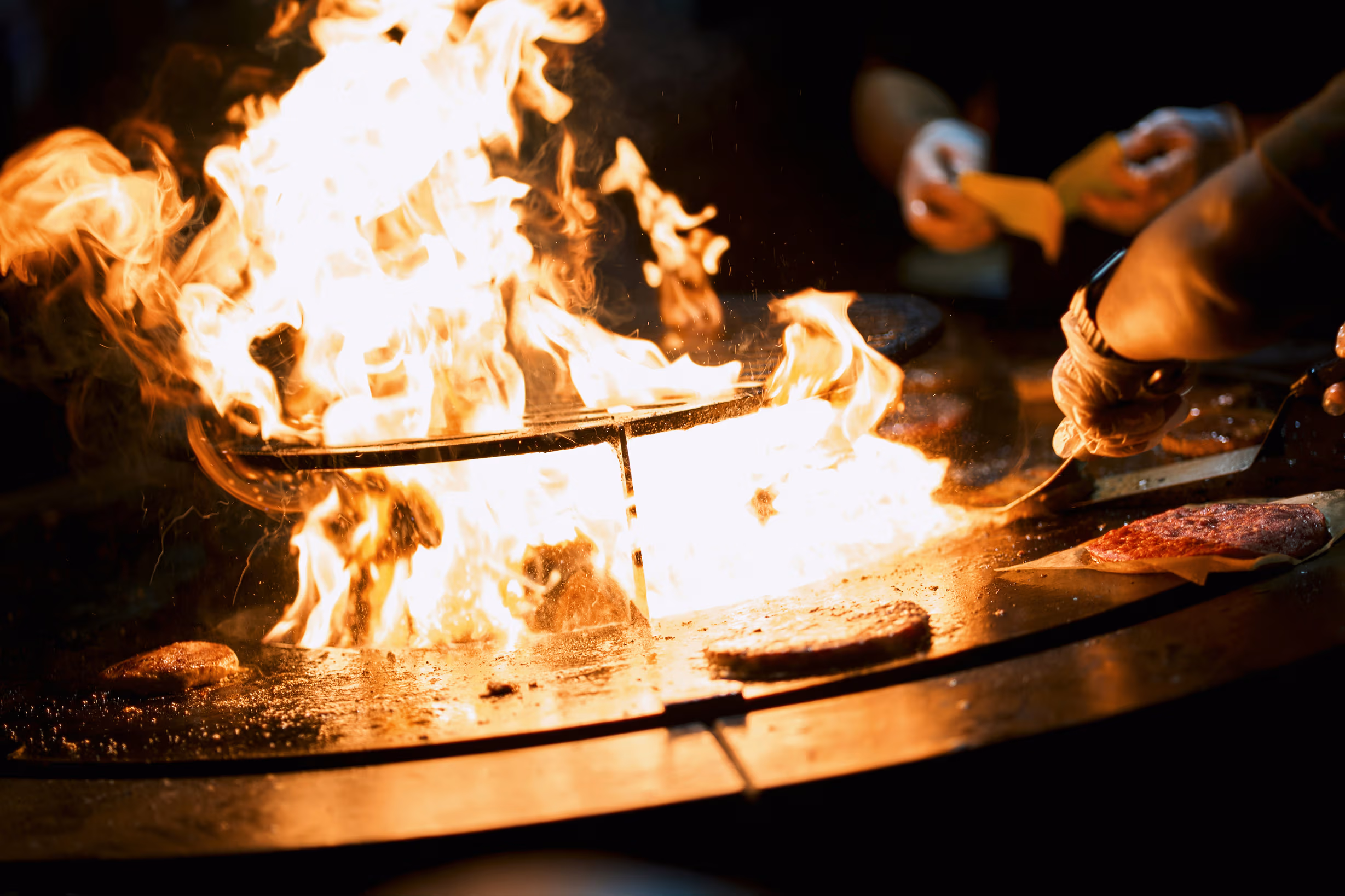 Patties on the round steel plate, flames rising from the fire at its centre.